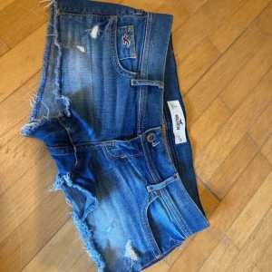 Blå jeansshorts från Hollister - Snygga blå jeansshorts från Hollister med råa, fransiga kanter och slitna detaljer. Klassisk femficksmodell med knapp och dragkedja framtill. Perfekta för varma dagar och ger en avslappnad vibe.