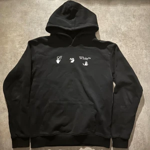 Svart Off-White hoodie med blått tryck - Säljer en svart hoodie från Off-White med vit logga och handmotiv på bröstet samt ett stort blått och vitt grafiskt tryck på ryggen. Klassisk känguruficka, huva och ribbade muddar. Perfekt för dig som gillar streetwear och vill sticka ut. Hör av vid minsta fundering 💫