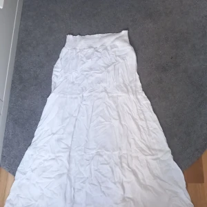 Vit långkjol från 157, stl M - Luftig och flowy vit långkjol från 157 med smockad midja för extra komfort. Kjolen har flera sömmar som ger en snygg volang-effekt och är perfekt för varma dagar. Enkel att matcha med crop tops eller stickade tröjor.