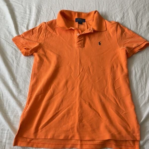 Orange pikétröja från Polo Ralph Lauren - Snygg orange pikétröja från Polo Ralph Lauren med klassisk krage och två knappar framtill. Tröjan har det ikoniska broderade logotypen i mörkblått på bröstet och är tillverkad i mjuk bomull. Perfekt för en chill och sportig look.