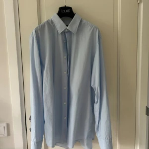 Ljusblå skjorta från Lauren - Klassisk ljusblå skjorta från Hugo Boss med lång ärm och knappar framtill. Skjortan har en rak passform och är tillverkad i mjuk bomull, perfekt för en clean och stilren look.