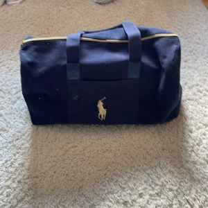 Mörkblå duffelbag från Polo Ralph Lauren - Snygg mörkblå duffelbag från Polo Ralph Lauren med klassisk guldfärgad logga framtill. Väskan har robusta handtag, dragkedja upptill och är gjord i tålig bomullscanvas. Perfekt storlek för en kort resa eller gymmet.