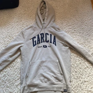 Beige hoodie från Garcia med tryck - Snygg beige hoodie från Garcia med mörkblå broderad logga och text på bröstet. Tröjan har huva med vita snören med svarta detaljer och en stor känguruficka framtill. Perfekt för chill dagar och enkel att matcha med jeans eller joggers.