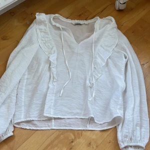 Vit blus med volang från Kappahl - Säljer en vit blus från Kappahl i storlek S. Blusen har långa ärmar med resår vid ärmslut, v-ringad hals med snörning och snygga volangdetaljer längs framsidan. Perfekt för dig som gillar romantisk och feminin stil.