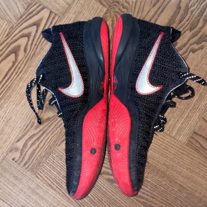 LeBron 20 - Säljer ett par svarta lebron  sneakers med stickad ovandel och stora vita Swoosh-loggor med röd kant. Skorna har snörning med svarta och vita mönstrade skosnören samt en riktigt fet röd yttersula med basketikon. Perfekt för dig som gillar sportig och unik stil.