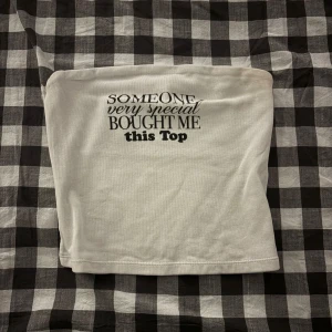 Vit bandeau tubtopp med text - Säljer en vit bandeau tubtopp med trycket 'Someone very special bought me this top' i svart. Toppen är axelbandslös och har en enkel, rak form som sitter tight mot kroppen. Perfekt för dig som gillar statement-plagg och minimalistisk stil.