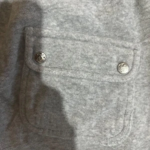 Grå mjukisbyxor från Juicy Couture - Snygga grå mjukisbyxor från Juicy Couture med bred resår och snörning i midjan. Byxorna har raka ben, stora fickor framtill med silverfärgade knappar och broderad JC-logga på ena fickan. Perfekta för chill eller träning.