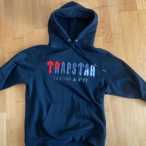 Trapstar hoodie - Svart hoodie från Trapstar med stor huva och snörning. Framsidan har ett fett tryck i rött, grått och blått med texten 'Trapstar It's a secret'. Tröjan har en klassisk känguruficka och långa ärmar. Perfekt för dig som gillar streetwear och vill sticka ut.