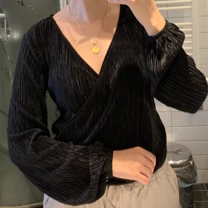 Svart omlottblus med plisserat material  - Säljer en svart omlottblus med plisserat material och långärmade, lite puffiga ärmar. Blusen har en djup v-ringning och är gjord i ett glansigt, mjukt material som ger en lyxig känsla. Perfekt för dig som gillar stilrena och trendiga plagg. Passar både xs och s 