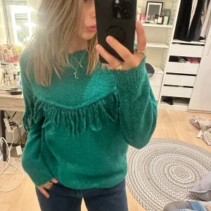 Grön stickad tröja med fransar - Snygg grön stickad tröja med fransdetaljer över bröstet. Modellen är långärmad och har en loose passform som ger en chill vibe. Perfekt att matcha med jeans för en cool look. Materialet är mjukt och känns skönt mot huden.