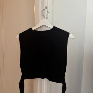 Svart croppad topp med knyt i ryggen - Snygg svart croppad topp med ärmlös design och cool öppen rygg som knyts längst ner. Perfekt att styla med höga jeans eller kjol för en trendig look. Materialet känns mjukt och stretchigt, vilket gör den bekväm att bära hela dagen.