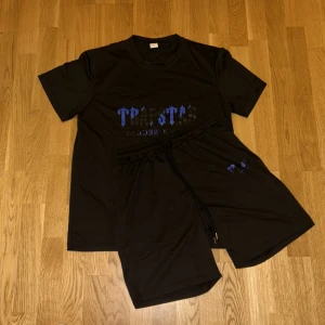 Svart t-shirt Trapstar med blått tryck - Svart t-shirt från Trapstar med blått tryck på bröstet där det står 'Trapstar It's a Secret' i gotisk stil. T-shirten har rund hals och korta ärmar, tillverkad i mjukt material som känns skönt mot huden. Perfekt för dig som gillar streetwear och vill sticka ut.