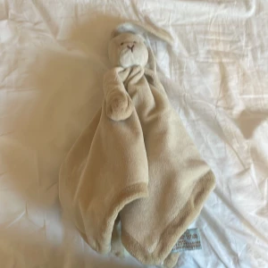  Snuttefilt  - Bilderna visar en mjuk beige snuttefilt med ett kaninhuvud och långa öron, från märket Diinglisar. Produkten är inte en bok, utan en gosedjursfilt som passar perfekt för mysiga stunder och trygghet. Perfekt för dig som gillar söta och mjuka accessoarer.