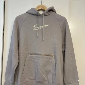 Grå Nike hoodie - Snygg grå hoodie från Nike i oversized fit med klassisk logga i vitt och guld på bröstet. Tröjan har huva med snörning, stor ficka med dragkedja och mjukt fleecefoder. Perfekt för chill dagar.