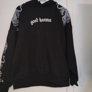 Svart hoodie med tigertryck från FB Sister - Svart hoodie med ljusgråa tigertryck på ärmar och axlar. På bröstet står det 'good karma' i ljusgrå text. Tröjan har huva och är långärmad, tillverkad i mjuk bomull och polyesterblandning. Stl M