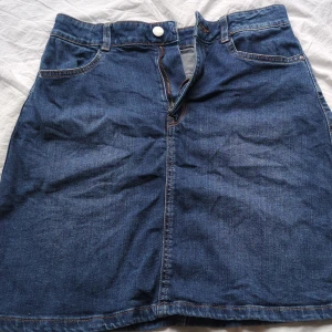 Mörkblå jeanskjol med fickor - Snygg mörkblå jeanskjol med klassisk A-linjeform och fyra fickor, inklusive två bakfickor och en framficka. Kjolen har hällor för bälte och stängs med dragkedja och knapp framtill. Perfekt för dig som gillar en enkel och cool denimstil.