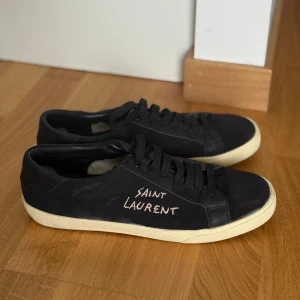 Svarta Saint Laurent sneakers - Svarta sneakers från Saint Laurent med vit text på sidan och vit sula. Skorna har snörning och en rund tå, med ovandel i canvas och detaljer i skinn. Snygg och stilren design som passar till många olika outfits. Skick 7.5/10. Välvårdade