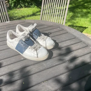Valentino Open sneakers vit/blå - Säljer ett par Valentino Open sneakers i vitt med breda blå paneler på sidorna. Skorna har snörning, rund tå och platt sula. Tillverkade i skinn med klassisk streetstil och diskret logga på sidan. Perfekta för dig som gillar lyxiga märken och clean design.