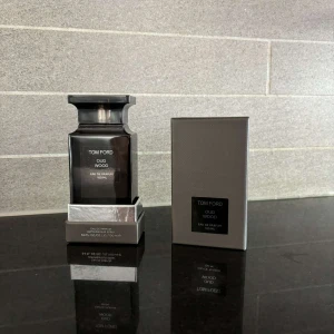 Tom Ford Oud Wood Eau de Parfum 100ml - Tom Ford Oud Wood Eau de Parfum 100ml med lyxig mörkgrå flaska och matchande kartong. Flaskan har en elegant, fyrkantig form med svart etikett och silverdetaljer. Oud Wood är känd för sin sofistikerade och träiga doftprofil som sticker ut.