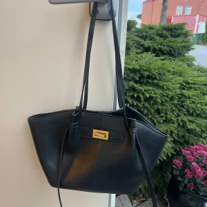 Svart handväska med gulddetalj - Snygg svart handväska med stilren form och långa axelband. Väskan har en elegant guldfärgad spänndetalj framtill och är tillverkad i slätt skinnmaterial. Perfekt för dig som gillar minimalistisk design och vill ha en väska som sticker ut med sin coola look.