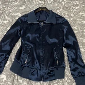 Mörkblå satin bomberjacka  - Snygg mörkblå bomberjacka i glansigt satinmaterial. Jackan har ribbade muddar, dragkedja framtill och två fickor med dragkedja. Perfekt för dig som vill ha en stilren och cool look med lite extra shine. Använd innan men inga problem vad jag har sett 