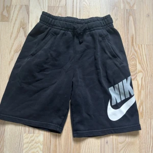 Svarta Nike shorts med logga - Svarta shorts från Nike med stor vit logga på ena benet. De har elastisk midja med snörning och två sidofickor. Mjukt material som känns skönt mot huden, perfekt för chill eller sport. Klassisk loose passform och enkel stil.