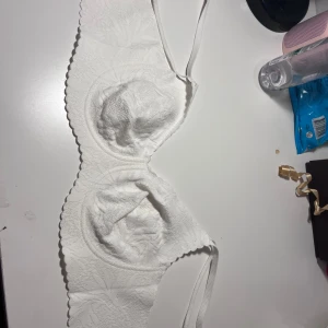 Vit BH med spetsdetaljer - Säljer en vit BH med snygga spetsdetaljer och blommigt mönster. Bikinin har en feminin form med scallop-kant och tunna axelband. Materialet känns mjukt och har en elegant struktur som ger en lyxig känsla. Perfekt för dig som vill ha en stilren och romantisk look.