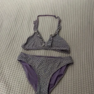 Ljuslila blommig bikini från Firefly - Supersöt ljuslila bikini från Firefly med små vita blommor över hela tyget. Toppen har volangdetaljer och halterneck med knyt i nacken. Materialet är mjukt och stretchigt, 84% polyamid och 16% elastan. Perfekt för strandhäng och poolparty! Den har också en liten lila rand på toppens band runt nacken