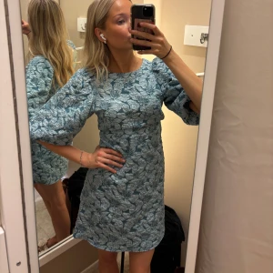 Blommig ljusblå kortklänning - Supersöt ljusblå kortklänning med blommigt mönster, lite skimrig och puffiga trekvartsärmar. Passar perfekt till bröllop! Helt oanvänd, lapp sitter kvar. Nypris 1200kr, från Fiore. Storlek 34 men passar även mig som brukar ha 36.