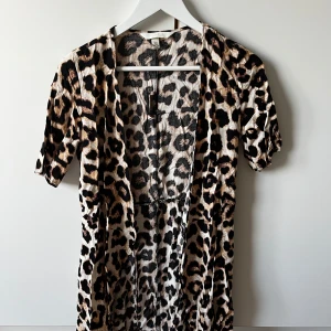 Leopardmönstrad omlottklänning från H&M - Säljer en snygg leopardmönstrad omlottklänning från H&M med korta ärmar och snörning i midjan. Klänningen har beige, svart och bruna toner och är gjord i mjuk polyester. Perfekt för dig som vill sticka ut med ett djuriskt print.