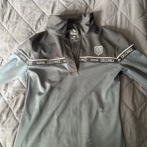 Svart ridtröja från Maya Delorez - Ridtröja från Maya Delorez med tryck och textdetaljer längs axlarna. Tröjan har lång ärm, krage och knappar framtill. Materialet är mjukt och stretchigt 💕 ursprungspris är 800 kr 💕