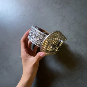 Glittrigt vitt bälte med stora nitar - Säljer ett brett vitt bälte med glittriga detaljer och stora silvriga nitar längs hela bandet. Spännet är dekorerat med gnistrande stenar och har en cool western-vibe. Perfekt accessoar för att lyfta vilken outfit som helst med extra bling.