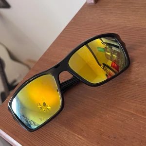 Svarta sportiga solglasögon med gula spegelglas - Säljer ett par svarta solglasögon med breda bågar och gula spegelglas. Glasögonen har en sportig design med röda detaljer vid gångjärnen. Perfekta för dig som gillar en cool och modern look. Ramen är i blank plast och glasen är rektangulära.