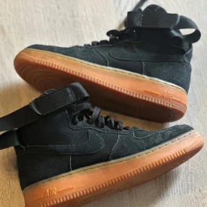 Nike Air Force 1 High svart mocka - Svarta Nike Air Force 1 High sneakers i mocka med högt skaft och klassisk snörning. Skorna har en snygg gummisula i orange ton och justerbar rem runt ankeln.
