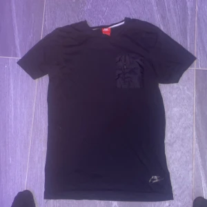 Svart Nike t-shirt med ficka - Svart t-shirt från Nike i storlek M. Klassisk rund hals och korta ärmar. På bröstet finns en snygg ficka med dragkedja som ger en sportig vibe. Materialet är mjuk bomull, perfekt för chill eller häng med kompisar.