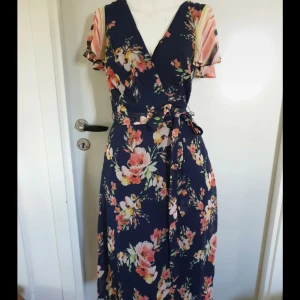 Blommig omlottklänning med knytband - Snygg långklänning i marinblått med blommigt mönster i rosa, vitt och gult. Klänningen har omlottmodell med v-ringning, kortärmade ärmar med färgglada ränder och ett knytband i midjan. 