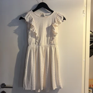 Vit kort klänning med volanger Kappahl - Supersöt vit klänning från Kappahl i storlek 146. Klänningen har broderade detaljer, volanger på axlarna och ett lätt utställt kjolparti. Perfekt för sommarens alla tillfällen! Materialet känns mjukt och luftigt, och den har rund halsringning.