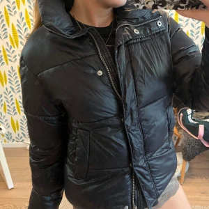 Svart pufferjacka - Snygg svart pufferjacka med två stora fickor framtill. Jackan har glansig yta, dragkedja och tryckknappar. Perfekt för kalla dagar och har en cool, lite boxig passform som ger streetkänsla.