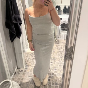 Glittrig silver långklänning med öppen rygg - Säljer en snygg silverfärgad långklänning med glittrigt tyg och tunna axelband. Klänningen har en tight passform och en riktigt cool öppen rygg med flera smala band som detalj. Perfekt för dig som vill sticka ut och glänsa på nästa event!