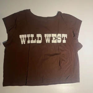 Brun croppad topp  - Brun croppad topp från H&M Divided med trycket 'WILD WEST' i vit, grov text på framsidan. Modellen har kortare till inga ärmar med rund hals. Sitter olika beroende på storlek. På mig (s-m) sitter den som en tajtare lite längre magtröja. Har bara används en gång så är i nyskick.