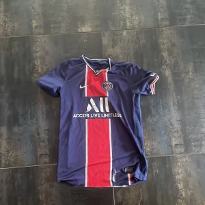 PSG matchtröja Nike blå/röd - Paris Saint-Germain fotbollströja från Nike i marinblått med bred röd vertikal rand och vita detaljer. Kortärmad med klubbmärke på bröstet och sponsortryck. V-ringad krage med vit och röd kant. Materialet är lätt polyester, perfekt för träning eller match.