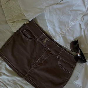 Brun jeanskjol med kontrastsömmar - Snygg brun mini jeanskjol med vita kontrastsömmar och klassiska fickor både fram och bak. Kjolen har bälteshällor och dubbla knappar i midjan. Perfekt att styla med en skjorta eller t-shirt för en trendig look. Storlek står ej med men passar mig som är vanlig xs-xxs