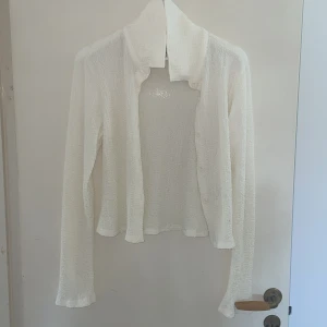 Vit kofta med krage och knappar - Säljer en vit kofta med lång ärm och klassisk krage. Koftan har en lätt textur och stängs med diskreta knappar framtill. Perfekt att slänga över en topp för en chill och clean look. Materialet känns mjukt och luftigt mot huden.