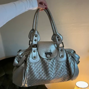 Silvrig handväska med flätad design - Snygg handväska i metallic silver med flätad struktur på framsidan och sidorna. Väskan har dubbla handtag, dekorativa nitar och spännen samt ett lock med metallspänne. Materialet är skinn och den har flera fack och detaljer som ger en cool vibe.