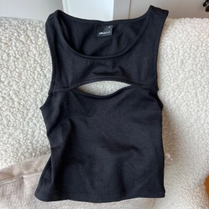 Svart ribbad croppad topp Gina Tricot - Svart croppad topp från Gina Tricot med ribbad struktur och snygg cut-out detalj framtill och baktill. Modellen är ärmlös och har en rundad halsringning. Perfekt för dig som gillar trendiga och stilrena plagg med en twist.