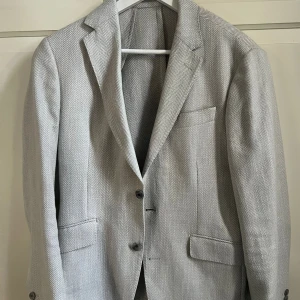 Beige kavaj från Hackett London - Snygg ljusgrå kavaj från Hackett London med diskret fiskbensmönster. Klassisk passform med två knappar framtill, fyra knappar vid ärmslut och flera fickor. Materialet känns lyxigt och är troligtvis ull eller linne, perfekt för en stilren look. En av knapparna har låsnat en gång med i tillbaka syd utan problem så de syns inte 