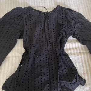 Svart blus med broderade hål från KappAhl - Svart blus från KappAhl med broderade hål och lång puffärm. Blusen har rund halsringning med knapp bak i nacken och är gjord i ett lätt, luftigt tyg. Perfekt för dig som gillar detaljer och vill ha en unik look.