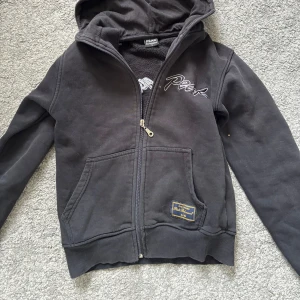 Svart hoodie från Peak Performance - Svart hoodie från Peak Performance med broderad logga både fram och bak. Tröjan har huva, dragkedja och två stora fickor framtill. Materialet är mjuk bomull och passformen är ganska liten.