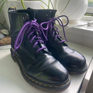 Svarta Dr. Martens boots med lila snören - Säljer ett par klassiska svarta Dr. Martens boots med lila snören och den ikoniska gula sömmen runt sulan. Skorna har rund tå, platt sula och är tillverkade i blankt skinn. Perfekt för dig som vill ha en edgy och tidlös stil. Är inte använda mycket, men har några repor som jag visar på bild