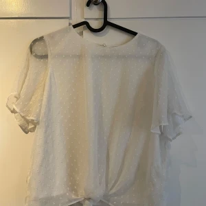 Vit prickig blus från Lindex - Säljer en söt vit blus från Lindex med diskret prickigt mönster och transparenta, luftiga korta ärmar. Blusen har en pärlknapp i nacken och en knytning framtill. Storleken är kids 158/164 sitter som xs/s